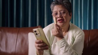 Görüntüler kilitli, gülümseyen Asya 'lı son sınıf kadını oturma odasındaki kanepede dinleniyor, cep telefonuna mutlulukla bakarken nefis atıştırmalıklar yiyor.