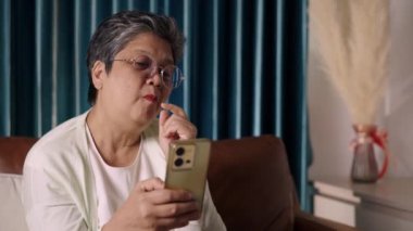 El kamerası görüntüsü, Asyalı neşeli yaşlı kadın oturma odasındaki koltukta akıllı telefondan görüntülü konuşma, cep telefonuna mutlulukla bakarken lezzetli atıştırmalıklar yediğini gösteriyor.