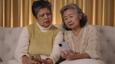 El kamerası görüntüsü, Front view, koltukta birlikte oturan iki yaşlı kadın, akıllı telefonlarına dikkatle bakıyorlar, oturma odasında sohbet ediyorlar ve arkadaşlıklarının mutlu anlarını paylaşıyorlar.