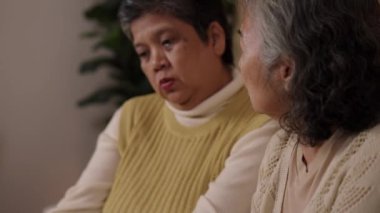 Yakın çekimde, iki yaşlı kadının yüz ifadesi, oturma odasındaki yakın ilişkide destek ve empati göstererek, akıllı telefona bakarken bir anlığına endişe duyuyor.