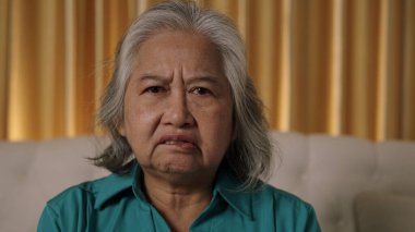 Gri saçlı yaşlı bir kadın yüzünde ciddi ya da kızgın ve düşünceli bir ifadeyle oturuyor. Bakışları doğrudan ve odaklanmış, derin düşüncelere ya da endişelere işaret ediyor..
