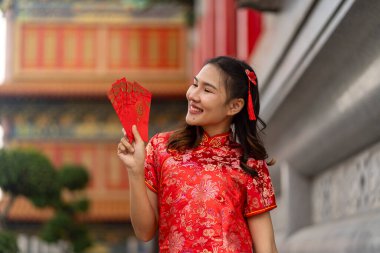 Canlı kırmızı Qipao gülüşü takınan ve geleneksel Çin tarzında dikilen genç bir kadın, elinde ay Çin Yeni Yılı 'nda iyi şans simgesi olan altın madalyalarla süslenmiş yelpaze kırmızı bir zarf tutuyor.