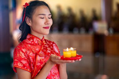 Portrait of Young woman In traditional red cheongsam Closed her eyes during holding  lit candle praying in temple on Chinese New Year, spiritual, cultural significance of Lunar New Year celebrations clipart