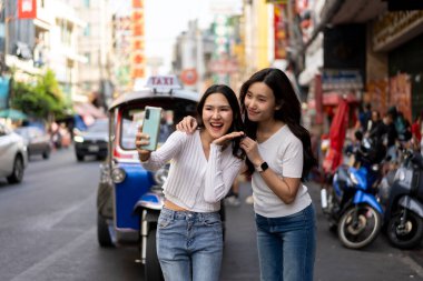 Genç kadın ve bayan arkadaş fotoğraf çekmek için akıllı telefonu kullanır, hareketli caddede renkli tuk-tuk ile mutlu poz verirler. Çin Mahallesi 'nde eğleniyorlar.