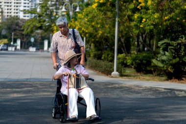 Asyalı yaşlı bir adam parkta karısını tekerlekli sandalyeye itiyor. Yeşilliklerle çevrili kıdemli çift boş boş gezmenin tadını çıkarıyor. Yaşlı çiftlerin parkında rahatlama. Boşluğu kopyala
