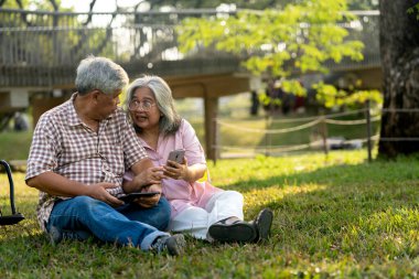 Asyalı yaşlı çift, halka açık parktaki dev ağacın altında güneşli ışıkta çimlerin üzerinde otururken cep telefonu kullanıyor. Yakınlarda park etmiş bir tekerlekli sandalye, akıllı telefona bakıyorlar ve birlikte mutlu bir şekilde gülümsüyorlar.