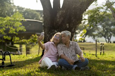 Asyalı yaşlı çift, halka açık parktaki dev ağacın altında güneşli ışıkta çimlerin üzerinde otururken cep telefonu kullanıyor. Yakınlarda park etmiş bir tekerlekli sandalye, akıllı telefon Selfie 'si kullanıyorlar ve birlikte mutlu bir şekilde gülümsüyorlar.