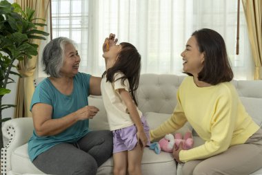 Samimi bir oturma odasındaki kanepede birlikte oynayan büyükanne, anne ve kızıyla bir aile bağının yürek ısıtan bir görüntüsü, sevgi ve beraberlik sergiliyor..