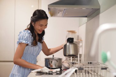 Genç Asyalı kadın modern mutfakta ev işi yapıyor, sobayı bezle temizliyor, günlük mavi elbiseler giyiyor, ev hayatı tarzında, temizlik ve hijyene odaklanıyor.