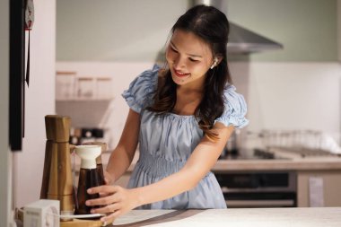 Genç Asyalı kadın mutfak tezgahını silerken gülümsüyor, açık mavi elbise, kulaklık takıyor, sıcacık ışıklar, düzenli modern mutfak, evdeki günlük yaşam tarzı