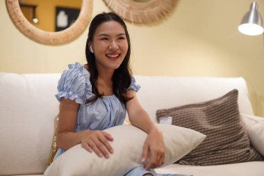 Açık mavi elbiseli genç Asyalı kadın kanepede oturuyor, yastığa yaslanıyor, nazikçe gülümsüyor, rahatlatıcı atmosfer, rahat iç mekan, huzurlu ve sakin ev hayatı sahnesi