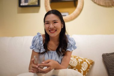 Güzel bir Asyalı kadın koltukta oturuyor, gülümsüyor ve elinde fincanla, rahat bir evde kulaklık takıyor yumuşak yastıklar ve sıcak aydınlatmalarla huzurlu ve neşeli bir anın tadını çıkarıyor.