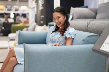 Sergi salonundaki açık mavi bir kanepede dinlenen genç Asyalı kadın, kolçaklara dokunurken memnuniyet içinde gülümsüyor, ev mobilyası alışverişi sırasında konfor ve yumuşaklık yaşıyor, gündelik ve zarif.