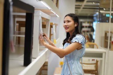 Genç Asyalı kadın mobilya mağazasında ahşap masayı keşfederken gülümsüyor, açık mavi elbise ve kablosuz kulaklık takıyor, alışveriş deneyiminden ve iç mekan ilhamından keyif alıyor.