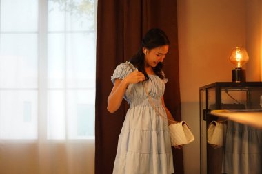 Açık mavi elbiseli genç Asyalı kadın gülümserken cam vitrinden beyaz el çantasını kaldırıyor, rahat ev dekorasyonu, sıcak ortam ışığı, şık bir yaşam tarzı.