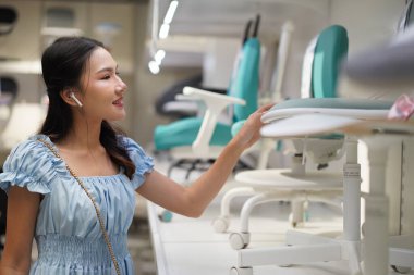 Mobilya mağazasında ofis başkanının yapısını ve konforunu değerlendiren Asyalı genç bir kadın, gülümseyen ve kulaklık takan, gündelik ve modern alışveriş deneyimi ve şık bir oturma tarzı var.