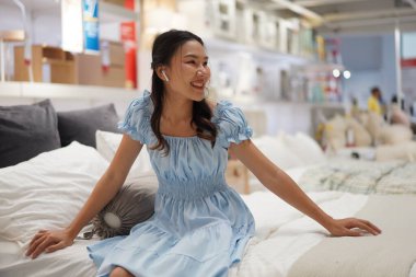 Açık mavi elbiseli genç Asyalı kadın modern mobilya mağazasında yumuşak bir yatağın tadını çıkarıyor, rahat ve mutlu hissediyor.
