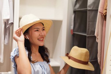 Genç Asyalı kadın gardıropta hasır şapka deniyor, farklı stilleri karşılaştırıyor, ev modası konsepti yumuşak ışıklandırma ve mutlu ruh hali.