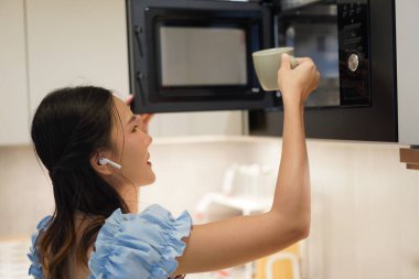 Modern ev mutfağında mikrodalga fırın kullanırken gülümseyen genç Asyalı kadın açık mavi elbise giyiyor, sıcak içeceği seramik bardakla hazırlıyor.