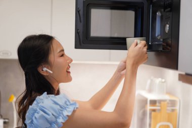 Asyalı bir kadın seramik bardak tutuyor, gülümsüyor ve mikrodalga fırına koyuyor rahat bir mutfakta, kablosuz kulaklıklarla ev hayatının tadını çıkarıyor.