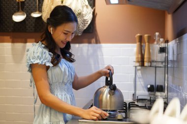 Genç Asyalı kadın, modern mutfakta ocakta metal çaydanlık kullanırken gülümsüyor, sıcak bir içecek hazırlıyor, açık mavi elbiseler giyiyor ve iç kısımları sıcacık.