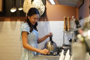 Uzun saçlı Asyalı kadın sıcak su hazırlıyor modern mutfakta çaydanlık kullanıyor, gülümsüyor ve açık mavi elbiseyle rahat vakit geçiriyor.