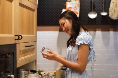 Hafif giysili Asyalı bir kadın sobada su ısıtıyor, gülümsüyor ve modern mutfak ortamında rahat vakit geçiriyor.