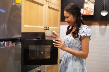 Mavi elbiseli genç Asyalı kadın içeceği ısıtmak için mikrodalga kullanıyor. Sıcak ev mutfağında mutlu bir şekilde gülümsüyor.