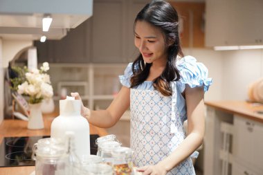 Beyaz seramik sürahi tutan önlük giymiş Asyalı bir kadın modern ev mutfağında yemek pişirmeye hazırlanıyor. Etrafı cam kavanozlarla çevrili.