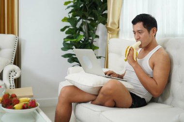 Asyalı adam evde beyaz bir kanepede muz yerken işine odaklanmış..