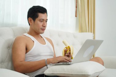 Asyalı adam dizüstü bilgisayar kullanıyor, elinde muz, evinde kanepede oturuyor..