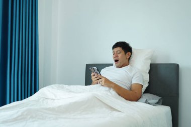 Uykulu Asyalı adam yatakta telefonu kontrol ederken esniyor. Yavaş ve tembel bir sabahı temsil ediyor..