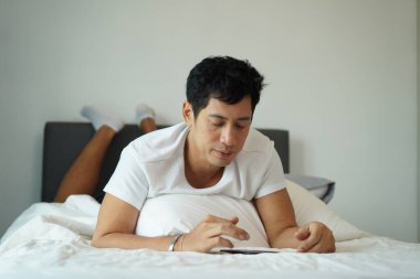 Asyalı bir adam sabah yatağında telefonuna göz atıyor, rahatlama ve dijital yaşam tarzını temsil ediyor..