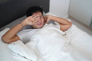 Parlak bir yatak odasında gözlerini ovuşturan Asyalı bir adam..