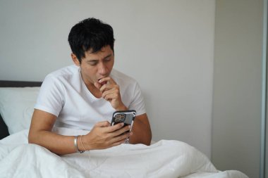 Yatakta yorgun bir adam akıllı telefon kullanırken esniyor, yorgunluk ve günlük telefon rutinini gösteriyor..