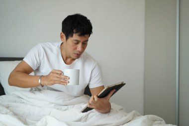 Beyaz tişörtlü Asyalı adam yatakta tablet okurken kahve içiyor. Sabah rahatlama konsepti.