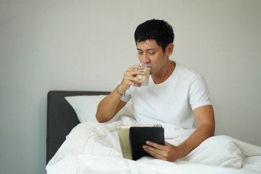 Genç Asyalı adam yatak odasında dinleniyor, soya sütü içiyor ve sabahları tablet okuyor.