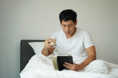 Beyaz tişörtlü rahat Asyalı adam soya sütü içip tablet tutuyor, yatak odasında huzurlu bir sabah geçiriyor, sağlıklı bir yaşam sürüyor.