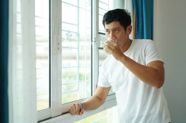 Genç Asyalı adam parlak pencerenin yanında soya sütü içiyor, günlük sağlıklı yaşam tarzı sabah sağlığı ve rahatlığını gösteriyor.
