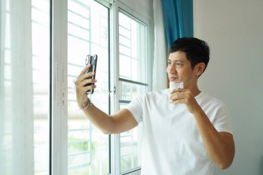 Soya sütü ve akıllı telefon taşıyan Asyalı adam gülümseyip selfie çekiyor ve günlük sağlıklı sabah rutininin tadını çıkarıyor.