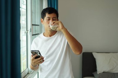Genç Asyalı adam pencereye yaslanmış telefon kullanıyor ve soya sütü içiyor. Huzurlu, modern bir sabah rutini ve sağlığını gösteriyor.