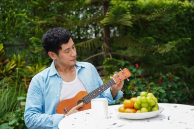 Asyalı bir adam bahçede beyaz masada oturmuş meyve tabağının yanında ukulele çalıyor ve huzurlu ve sağlıklı bir anın tadını çıkarıyor.