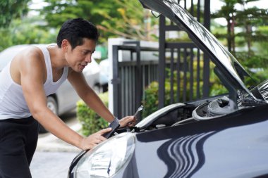 Asyalı bir adam İngiliz anahtarı ve el feneri tutuyor. Evin önünde arabanın kaputunu kontrol ediyor. Konsantrasyon ve sabırla aracın tamirini yapıyor.