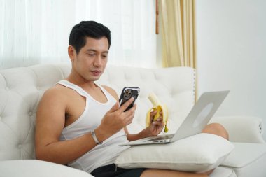 Asyalı adam elinde muz tutarken akıllı telefon kullanıyor ve evde dizüstü bilgisayarla oturuyor..