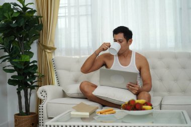 Asyalı adam kanepede bağdaş kurup kahve içiyor ve evde dizüstü bilgisayar kullanıyor..