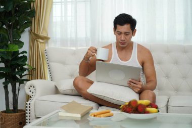 Evde rahat bir koltukta dizüstü bilgisayarla çalışırken kahvesini yudumlayan odaklanmış bir adam..