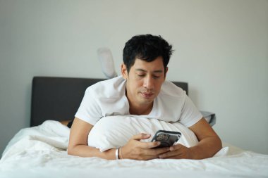 Rahat bir Asyalı sabah yatak odasında akıllı telefon kullanıyor..
