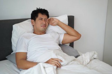 Asyalı bir adam yatakta oturur ve sabah yorgun ve uykulu bir ifadeyle gözlerini ovuşturur..