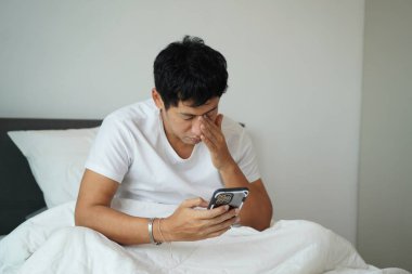 Uykulu adam gözlerini ovalarken akıllı telefonu kontrol ediyor. Sabah dijital alışkanlığı ve yorgunluk kavramı.