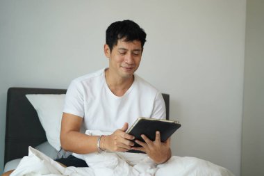 Gülen adam yatakta oturuyor ve sabahları dijital tablet kullanıyor. Pozitif ruh hali ve rahatlamış sabah rutini.